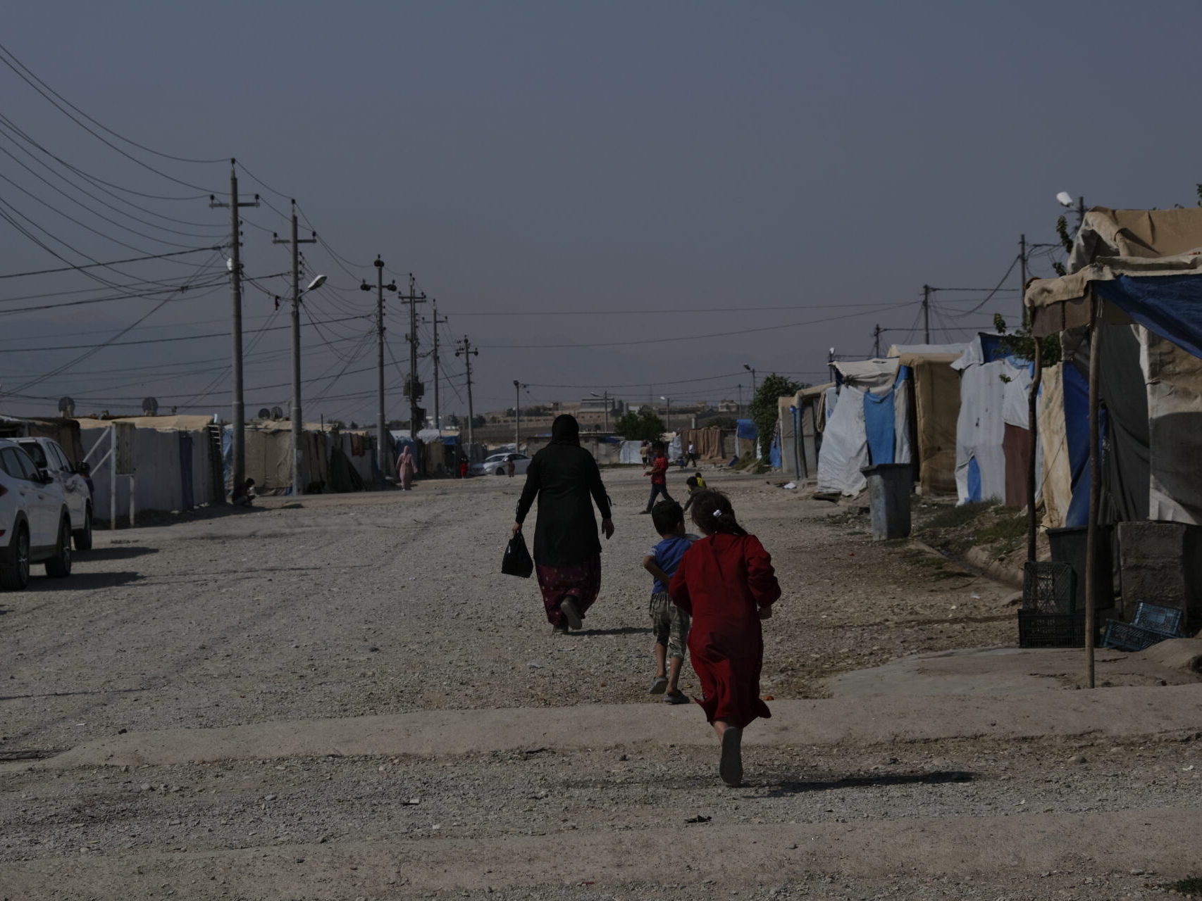 Für Frieden im Irak. Auf dem Foto zu sehen: Eine Frau und mehrere Kinder laufen auf einem breiten Kiesweg in einem Camp aus notdürftig zusammengezimmerten Unterkünften. Die Straße wird gesäumt durch Stromleitungen und ein paar Autos.