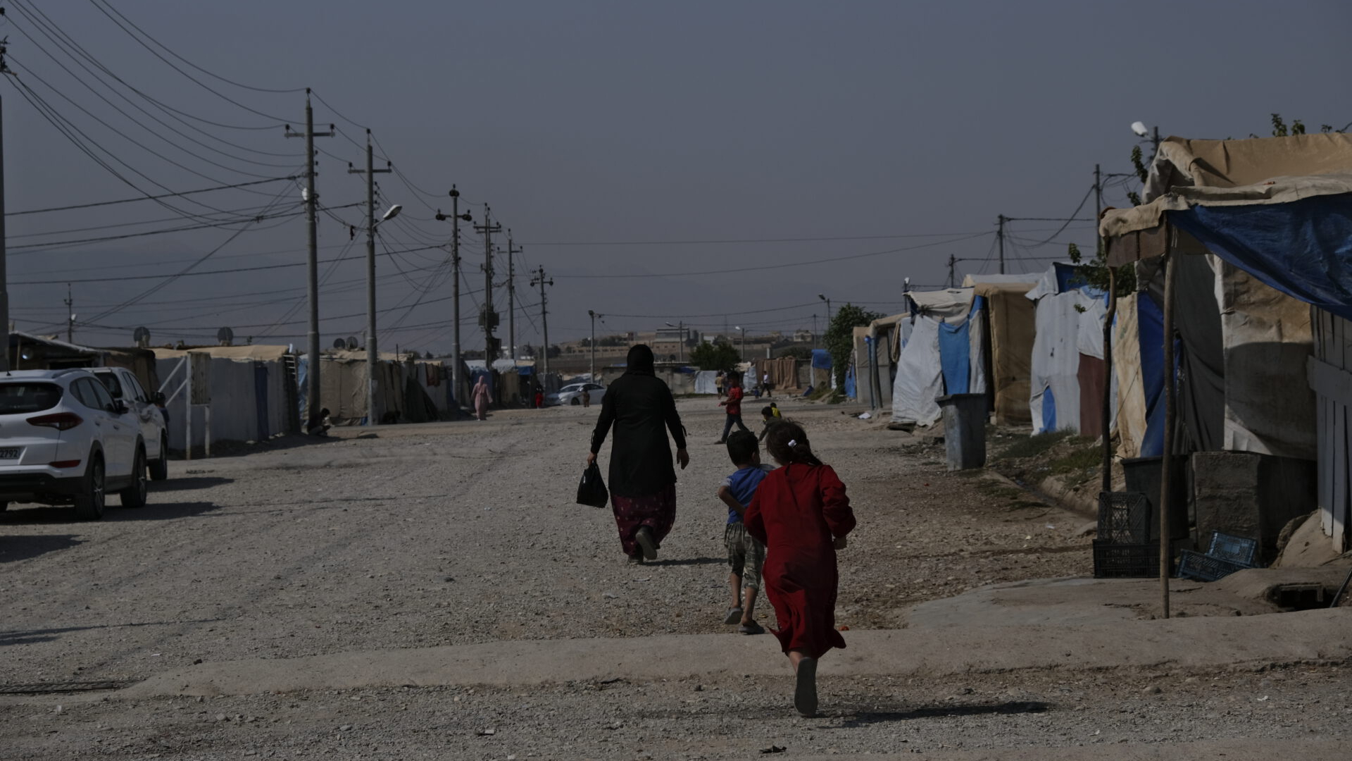 Für Frieden im Irak. Auf dem Foto zu sehen: Eine Frau und mehrere Kinder laufen auf einem breiten Kiesweg in einem Camp aus notdürftig zusammengezimmerten Unterkünften. Die Straße wird gesäumt durch Stromleitungen und ein paar Autos.