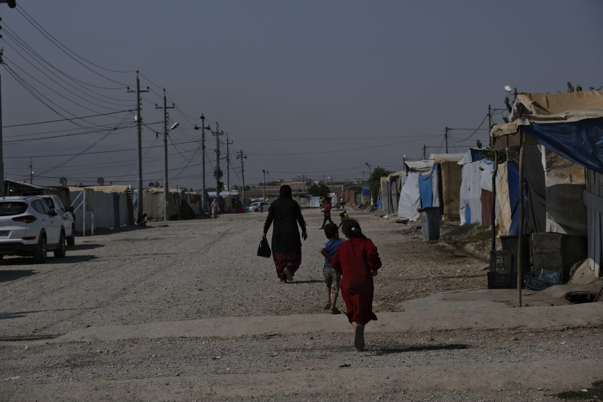 Für Frieden im Irak. Auf dem Foto zu sehen: Eine Frau und mehrere Kinder laufen auf einem breiten Kiesweg in einem Camp aus notdürftig zusammengezimmerten Unterkünften. Die Straße wird gesäumt durch Stromleitungen und ein paar Autos.